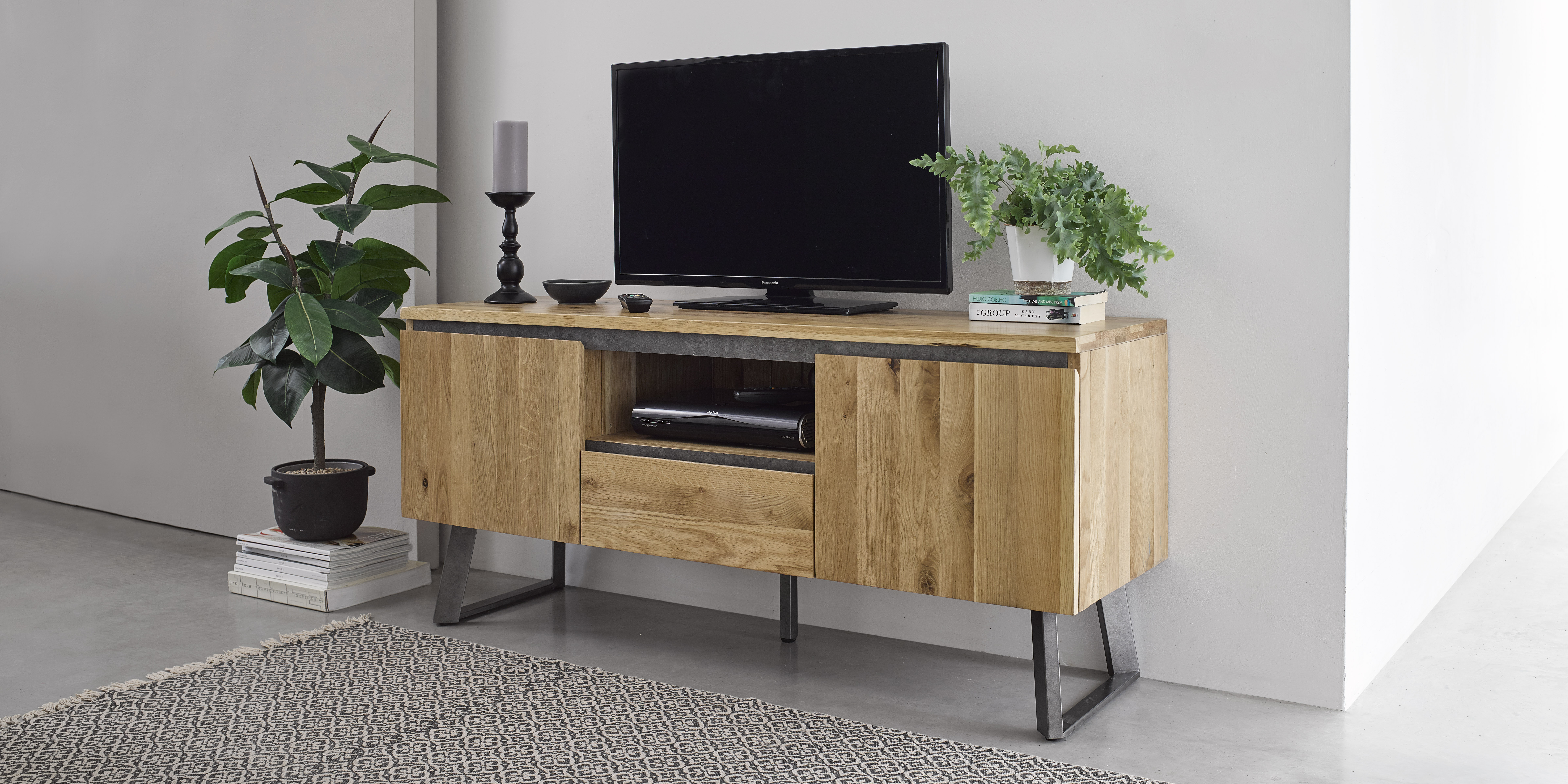 A light wood industrial style TV stand with a TV on in a light room with light grey walls and a light grey patterned rug. 